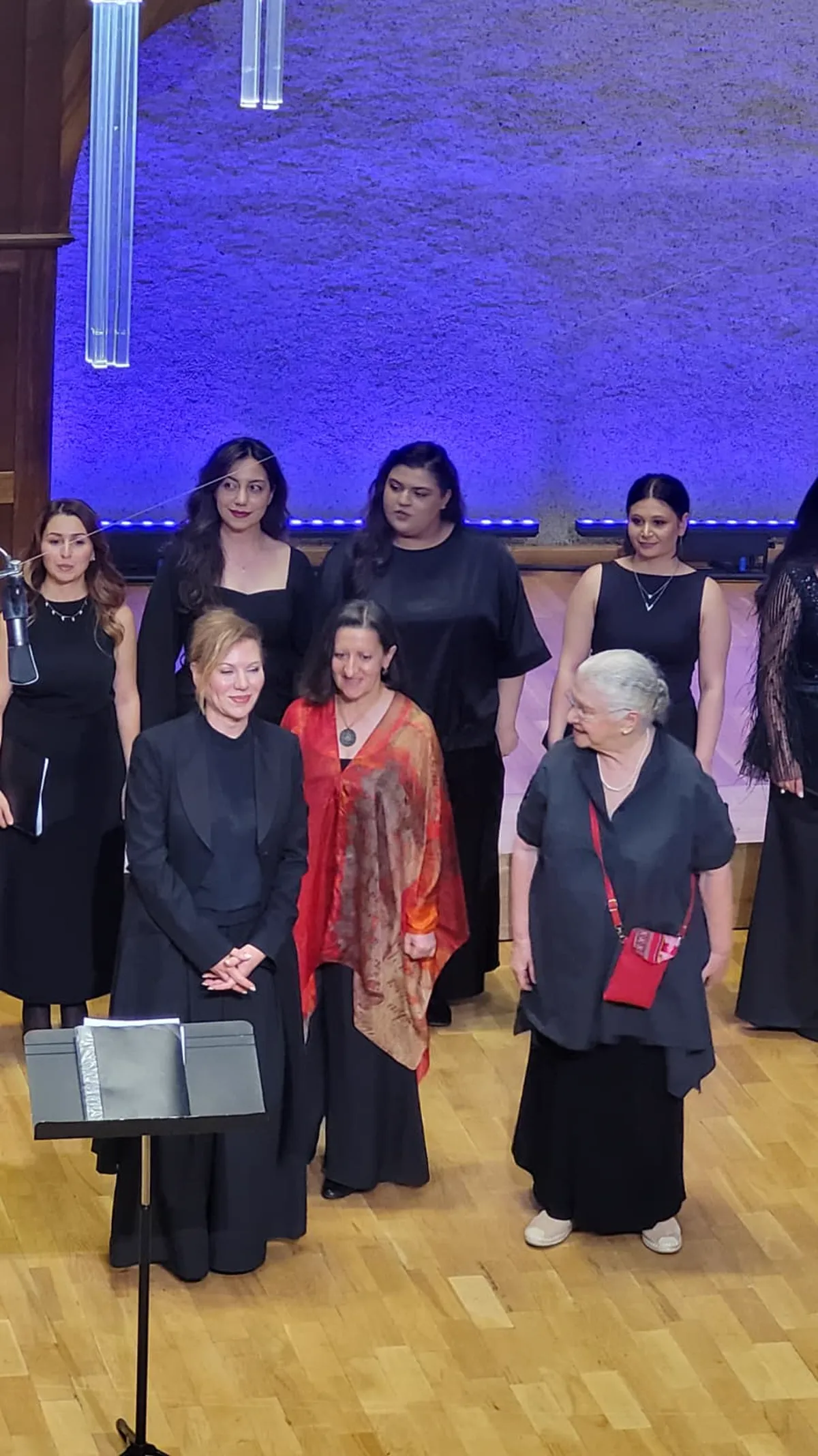 The choir and its conductor standing on a wooden stage under blue concert lighting